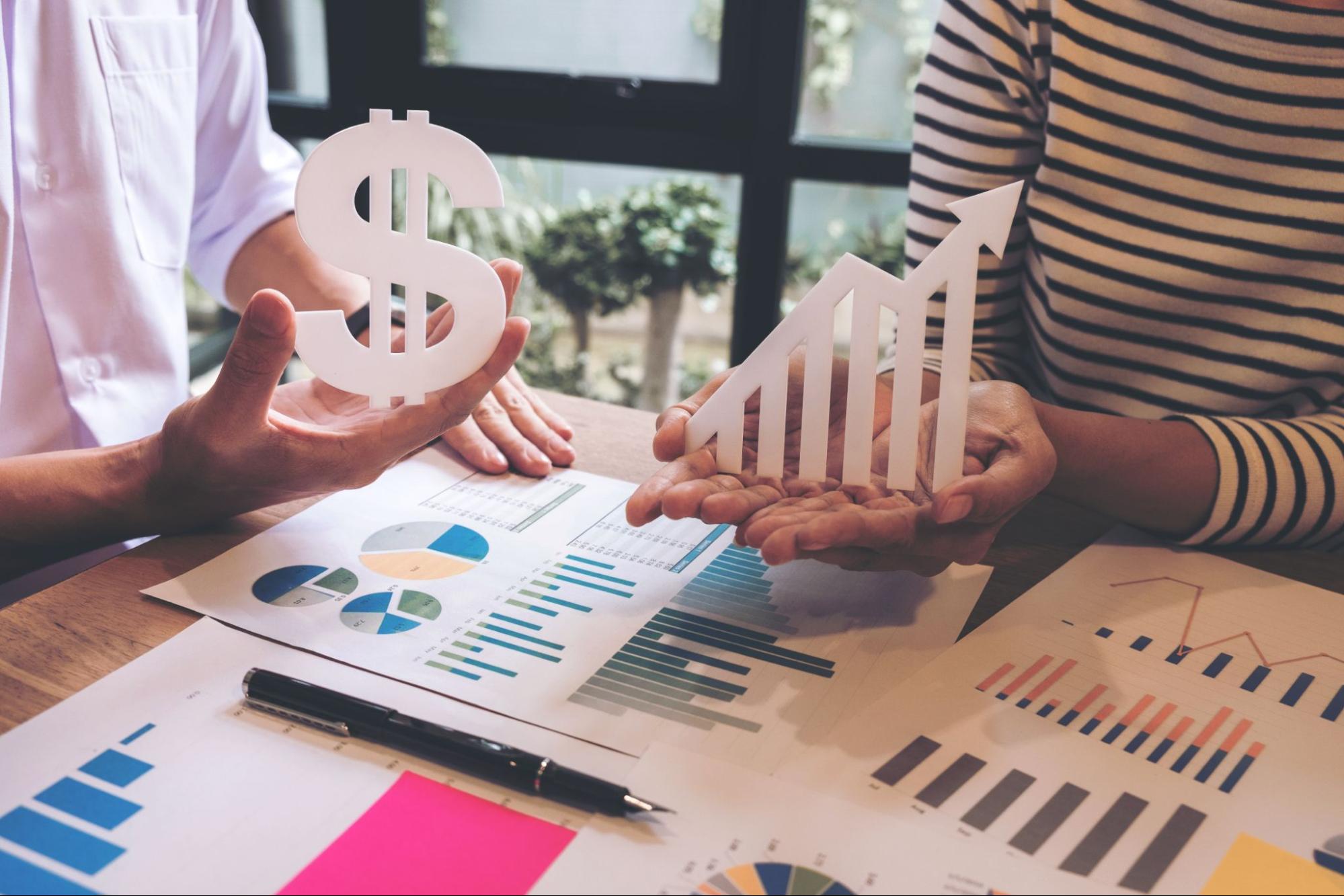 Two businessmen holding dollar and growth signs discuss budgeting plans, with various graphs and charts on the table illustrating budgeting basics.