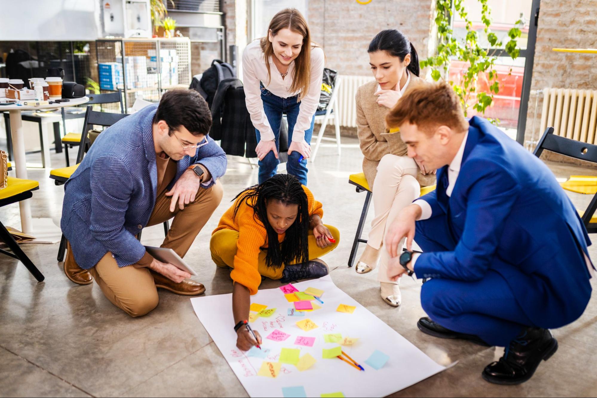 A business team uses sticky notes on a large white cartolina on the floor to brainstorm and develop a profitable business idea.
